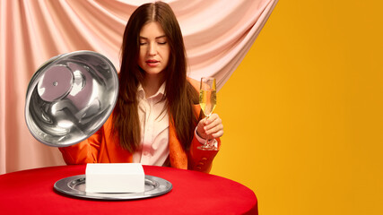 Woman with champagne and cloche revealing white copy space box on red table with pink drapery....