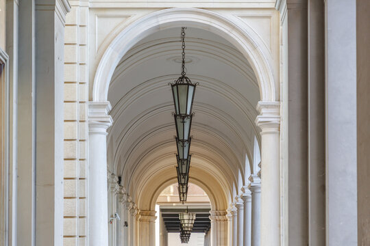 View of a long corridor, defined by light-colored arches and columns, with hanging lanterns casting soft shadows in a symmetrical display, Chieti, Abruzzo, Italy.