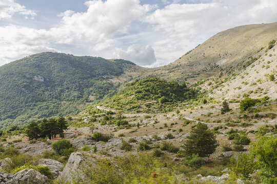 View of sunlight paints the rocky terrain and winding road through the lush, green, tree-covered hills under a vast, cloudy sky, Frattura Vecchia, Abruzzo, Italy.