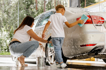 Happy mother and son washing a white car together with sponges and foam at self-service car wash, bonding activity on a summer day