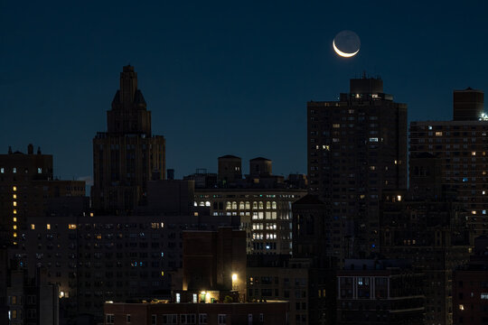 Fototapeta View of a crescent moon hanging above the dark, silhouetted buildings, glowing with scattered lights against the night sky, Moon in New York, New York, United States.