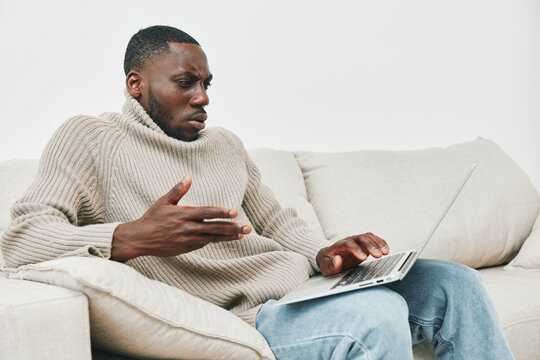 A thoughtful African American man in a cozy beige sweater sitting on a couch, engaging with a laptop, embodying curiosity and contemplation in a minimalist living space - Powered by Adobe