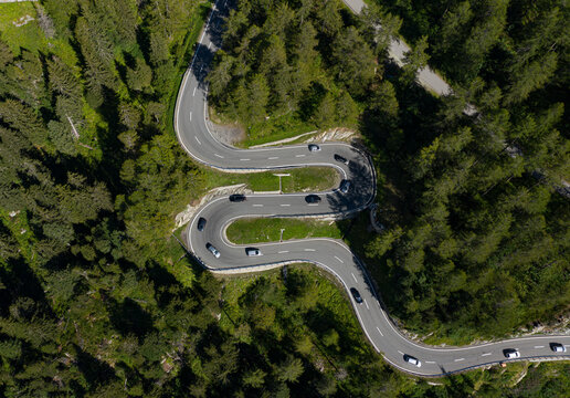 Aerial view of a serpentine road winding through a dense forest, cars navigating the sharp bends, contrasting with the deep green foliage, Maloja Pass, Maloja, Switzerland.