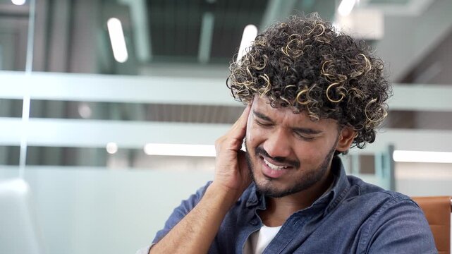 Sick young businessman suffers from ear pain while sitting in office. Upset male touches his ear with his fingers. He has furuncle, shingles, eczema, ear diseases or otitis media. Earache. Close up