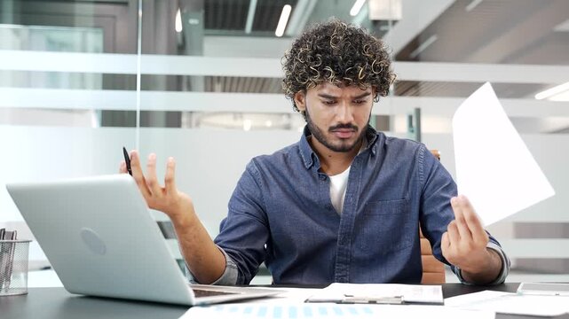 Confused puzzled young businessman having difficulty with paperwork sitting at workplace in business office. Frustrated financier reviewing documents on a laptop, unhappy with bad financial results