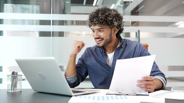 A smiling businessman is satisfied with results of a financial report looking at documents and a laptop screen sitting at workplace in a business office. Entrepreneur is happy with positive indicators