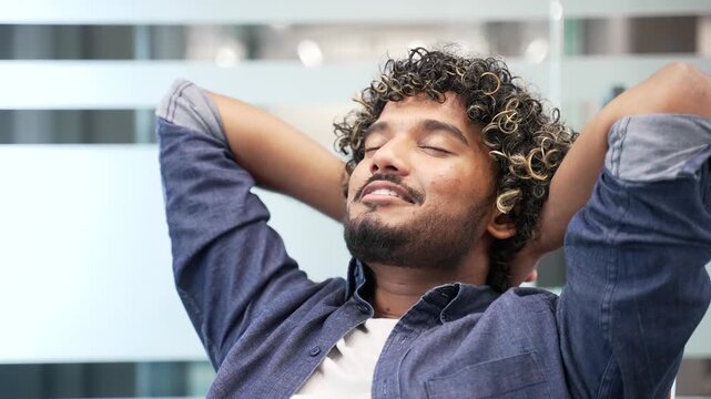 Happy young businessman resting puts his hands behind his head sitting on at workplace in business office. Smiling satisfied handsome male relaxes with closed eyes during break at work. Close up