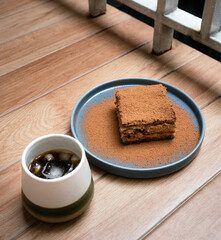 The photo shows a piece of tiramisu dessert on a blue plate, dusted with cocoa powder. Beside it, there is a cup of iced coffee placed on a wooden table near a balcony railing, creating a calm and coz