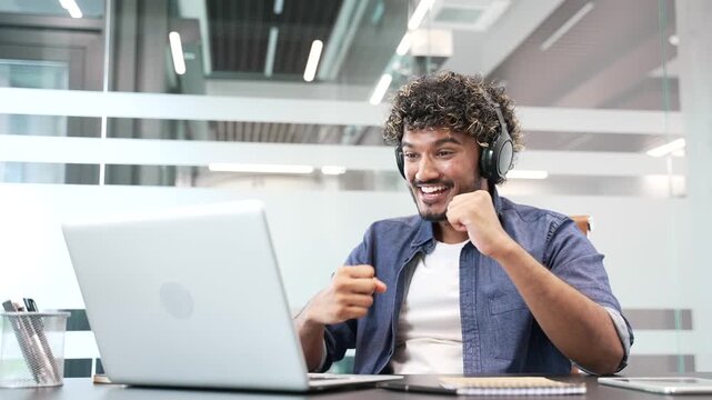 Excited joyful young businessman in headphones watching sports match or competition using laptop sitting at workplace in business office. Happy man cheering for bids at auction, celebrating success