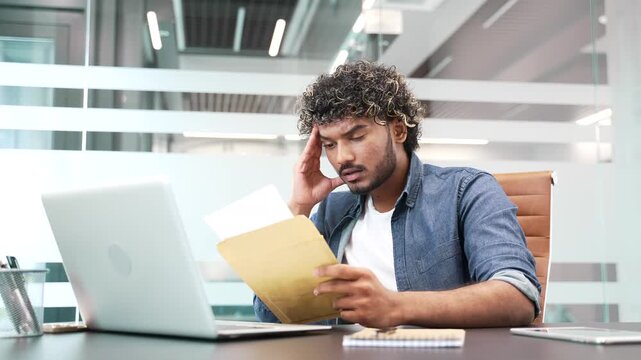 Upset disappointed businessman reading a letter with bad news sitting in a business office. Worried entrepreneur frustrated by receiving unpleasant negative notification after opening the envelope
