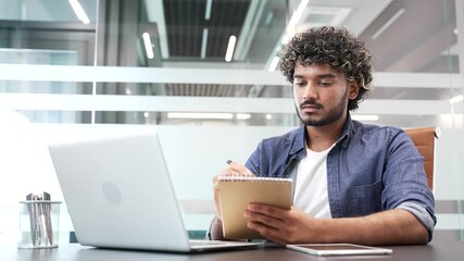 Young businessman watching video call conference or training takes notes looking at laptop screen sitting at workplace in business office. Focused worker listening to remote online course, seminar - Powered by Adobe