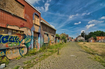 Wide angle view of deserted cobblestone street lined with derelict buildings covered in colorful graffiti under blue sky with clouds in abandoned Belgian village of Doel.