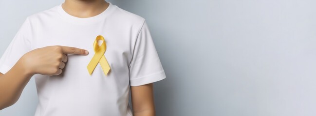 Caucasian boy stands with a yellow ribbon on a white t-shirt, pointing at it. Banner template for childhood cancer awareness month concept and support for disease patients with copy space
