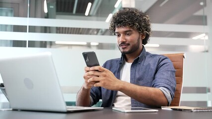 Smiling businessman is using browsing mobile phone while sitting at workplace in a modern business office. Handsome worker is chatting online, working in app or reading writing message on smartphone - Powered by Adobe