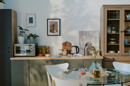 Modern kitchen interior featuring countertop with electric oven, glass jars, electric kettle, cutting board, and decorative plants, glass dining table with chairs in foreground