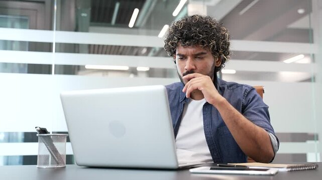 Upset frustrated businessman reads bad news on laptop while sitting at desk at workplace in modern business office. Worried male worker is sad while reviewing negative message information on computer