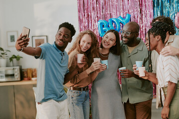 Group of diverse young adults and one pregnant young adult woman smiling and posing for selfie at party, holding cups and standing together in front of colorful backdrop with decorations
