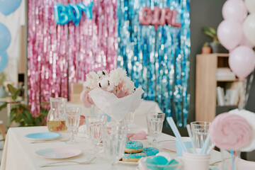 Table set with glassware, plates, donuts, and pink decorations standing in front of shimmering blue and pink backdrop with boy and girl signs, suggesting gender reveal party celebration