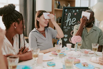 Group of young adults and Black man playing guessing game at party, holding cards to foreheads while sitting at table with desserts and drinks, smiling and interacting together