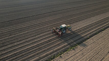 A modern tractor with agricultural equipment works across a vast plowed field creating perfect rows under a bright sky indicating a new growing season