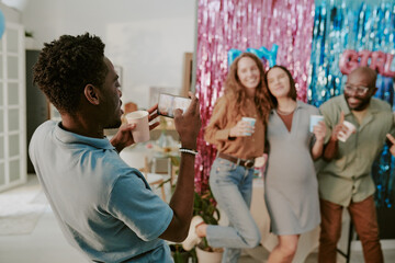 Young Black man photographing three smiling young adults including pregnant Caucasian woman and diverse friends holding cups standing together 