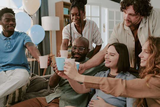 Diverse group of young adults and middle aged people smiling and raising paper cups in toast, multiethnic friends gathering together on couch celebrating special occasion indoors
