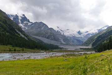 Roseg Valley and Bernina Glaciers. Swiss mountain valley with a glacial river, green meadows, snow-capped peaks under a dramatic sky. Alpine landscape of Engadin, Switzerland.
