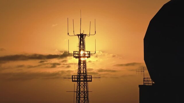 Telecommunication tower and parabolic antenna silhouette against vibrant orange sunset sky, essence of communication and technology in a breathtaking evening landscape. Aerial drone flight panorama