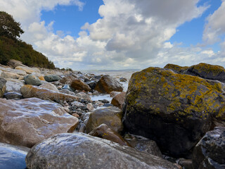 Shoreline with wet stones and lichen patches by the sea under cloudy sky, natural coastal landscape scene.