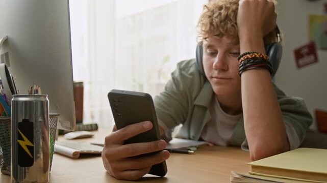 Close up shot of bored young man using smartphone while studying and lying on desk at home with focus flowing from face to phone