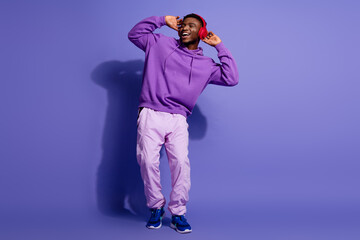 Young man wearing stylish purple hoodie enjoying music with headphones and smiling against a violet background