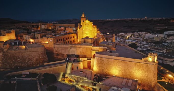Malta, Comino island: Majestically illuminated historic Citadel in Gozo. The ancient fortifications glow beautifully in the night electric lights. Aerial view drone flight footage rotation panorama