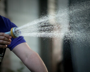 A person's arm holds a hose nozzle, spraying a wide stream of water outwards, creating a misty...
