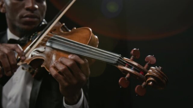 Focused young adult Black musician turning tuning pegs on violin preparing string instrument before music performance on stage against black background