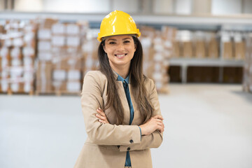A confident woman wearing a yellow hard hat and a blazer stands in a warehouse, showcasing leadership and professionalism.