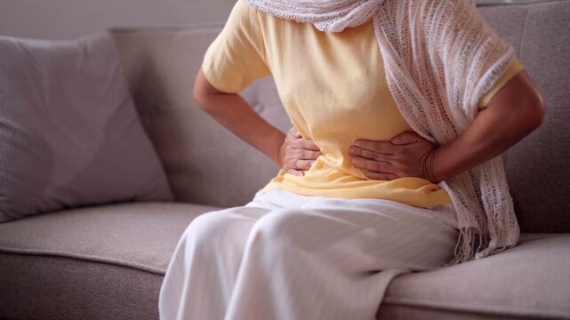 Older Asian woman experiences severe abdominal pain at home sofa health issue medical discomfort stomach ache
