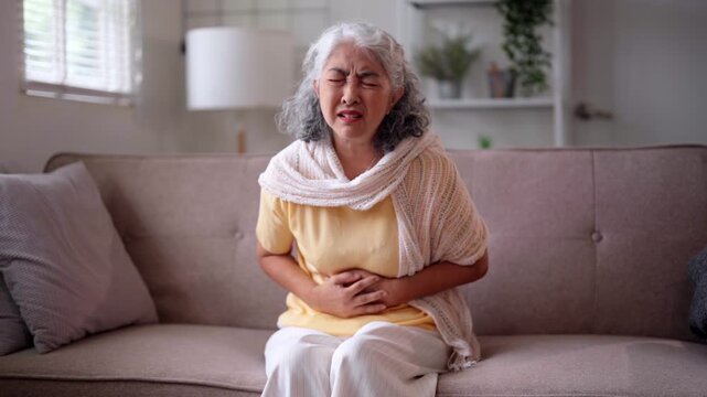 Older Asian woman sit sofa feel severe stomach pain. Abdominal discomfort and illness cause distress. She appear unwell suffer chronic health trouble at home.