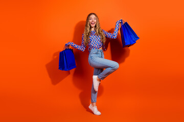 Energetic young female holding blue shopping bags smiling and posing against a vibrant orange background in fashionable attire