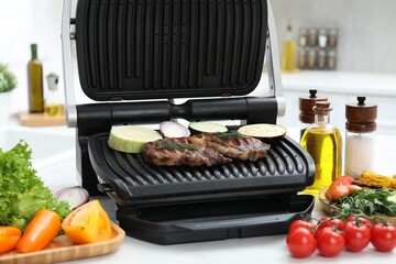 Electric grill with tasty meat, vegetables and spices on white table in kitchen