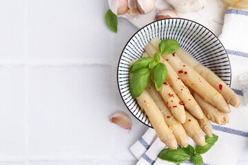 Pickled asparagus with spices, basil and garlic on white tiled table, flat lay. Space for text