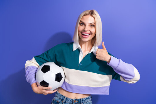 Blonde woman in casual colorful outfit holding soccer ball gesturing thumbs-up on purple background promoting sports