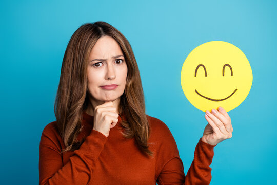 Portrait of a thoughtful woman holding a smiley face icon against a bright blue background