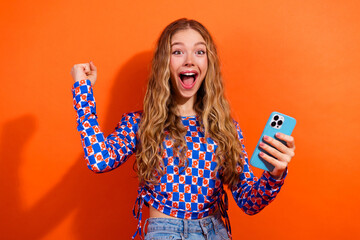 Happy young woman holding a smartphone celebrating and expressing excitement against a bright orange background