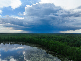 A panoramic aerial shot of a large, dark storm cloud covering a part of the sky over a dense green forest and a lake.