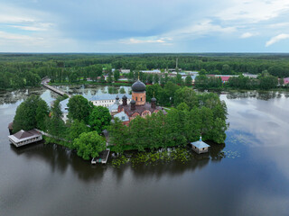 An aerial view of the Vvedensky Island Monastery, showing its red brick church and white buildings on a small island surrounded by a lake with lily pads.