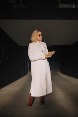 Stylish woman wearing a white coat and sunglasses stands in urban alleyway during daytime