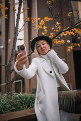 Woman in white coat takes a selfie in autumn park with yellow leaves