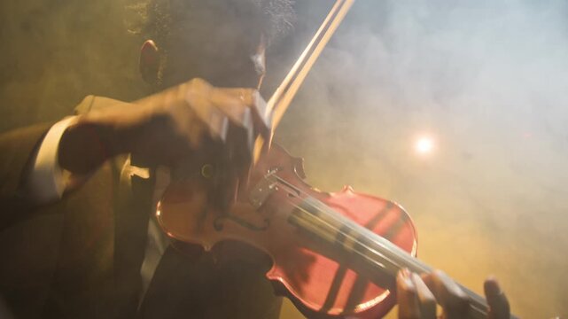 Young adult African American male violinist playing music on stage illuminated by spotlight and filled with mist during symphonic orchestra concert