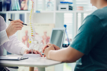A doctor is giving advice to a patient with back pain in a clinic using a skeleton model, laptop and whiteboard to record the patient's symptoms. Concept of healthcare and medical care.