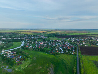 Fototapeta premium A wide aerial shot of the city of Suzdal, showing a mix of residential houses and a road, surrounded by vast green fields and forests.